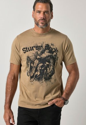 Mand med skæg iført en beige T-shirt med en stor grafik af et vædderkranium og tekst, med sorte bukser og et armbåndsur på venstre håndled.