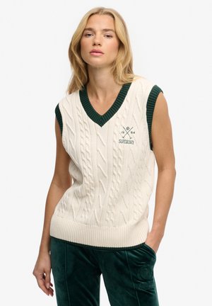 Femme aux cheveux blonds portant un pull sans manches blanc en maille torsadée avec des bordures vert foncé et un pantalon en velours vert foncé, debout, une main dans la poche.