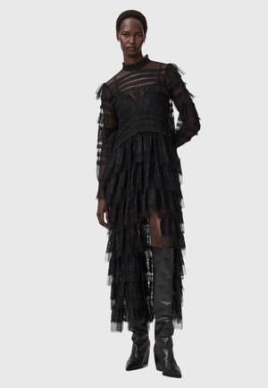 Svart spetsklänning med lager av rufser, långa genomskinliga ärmar och hög halsringning. Matchad med knähöga svarta stövlar, vilket framhäver de genomskinliga lagren.