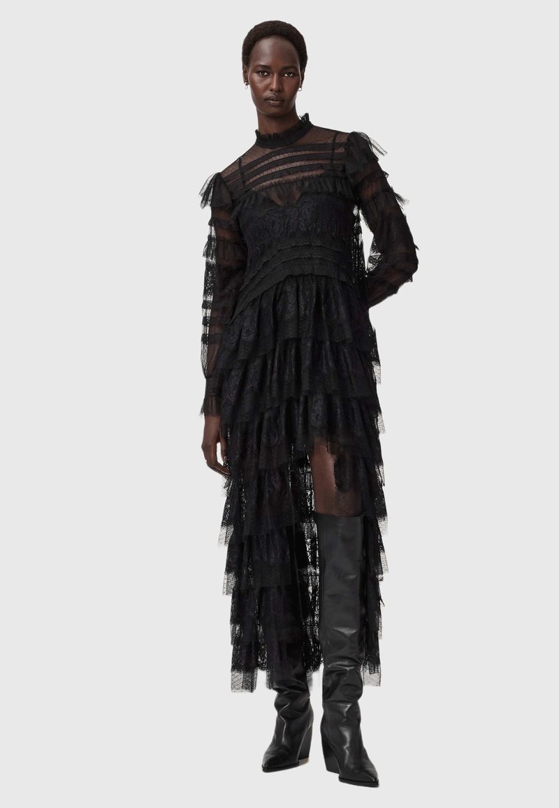 Svart spetsklänning med lager av rufser, långa genomskinliga ärmar och hög halsringning. Matchad med knähöga svarta stövlar, vilket framhäver de genomskinliga lagren.