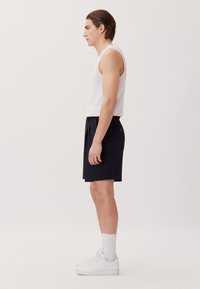 Top senza maniche di tessuto bianco con pantaloni corti neri su misura, dotati di tasca laterale e sneaker bianche con cuciture in evidenza.