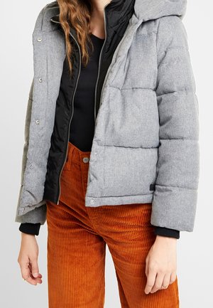 Kobieta w szarej pikowanej kurtce z czarną podszewką, czarnym topem i pomarańczowych, wysokowysokich spodniach sztruksowych, stoi na tle jednolitego tła.