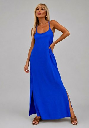 Femme aux longs cheveux blonds portant une robe maxi bleu roi sans manches avec des fentes latérales et des sandales marron sur un fond gris uni.