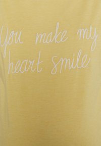 T-shirt de algodão amarelo com texto em branquíssimo cursivo que diz: "Você faz meu coração sorrir." Textura suave com um acabamento liso.
