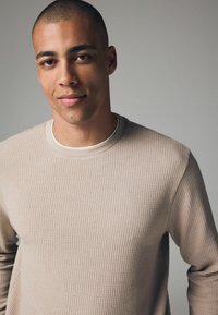 Suéter texturizado beige claro con cuello redondo y mangas largas, que presenta un sutil patrón de canalé y un acabado de tela suave.