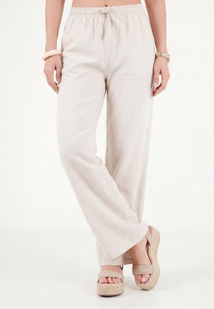 Femme portant un pantalon beige à jambes larges avec une ceinture élastique et des sandales plateformes beiges, debout avec une jambe croisée sur l'autre.
