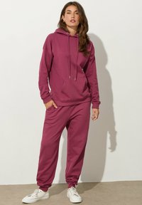 Hoodie color berry con lacci e tasca frontale, abbinato a pantaloni della tuta a vestibilità ampia. Presenta polsini a costine e tasche laterali.