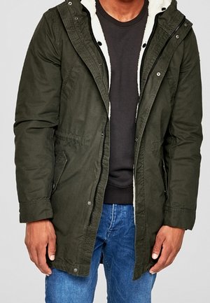 Homme portant un parka vert foncé avec une doublure en polaire blanche, un sweat-shirt noir et un jean bleu, debout avec les bras détendus le long du corps.