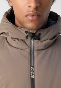 Chaqueta marrón con capucha y cuello alto. Presenta un cierre con cremallera y acentos a rayas en contraste. Tejido texturizado y herrajes ajustables con cordón.