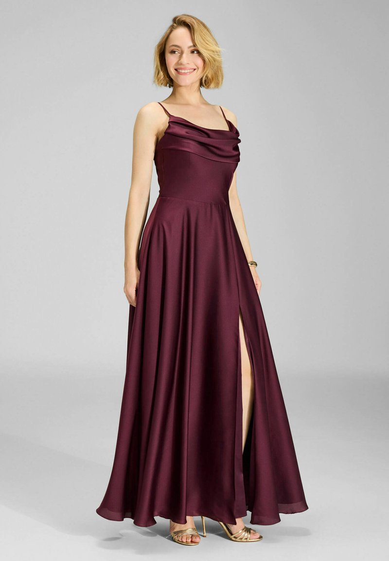 Femme souriante, portant une robe en satin violet foncé sans manches avec une fente latérale et des sandales à talons hauts dorées, debout devant un fond gris.
