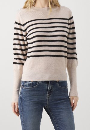 Femme portant un pull côtelé beige avec des rayures horizontales noires associé à un jean en denim bleu, debout sur un fond uni.
