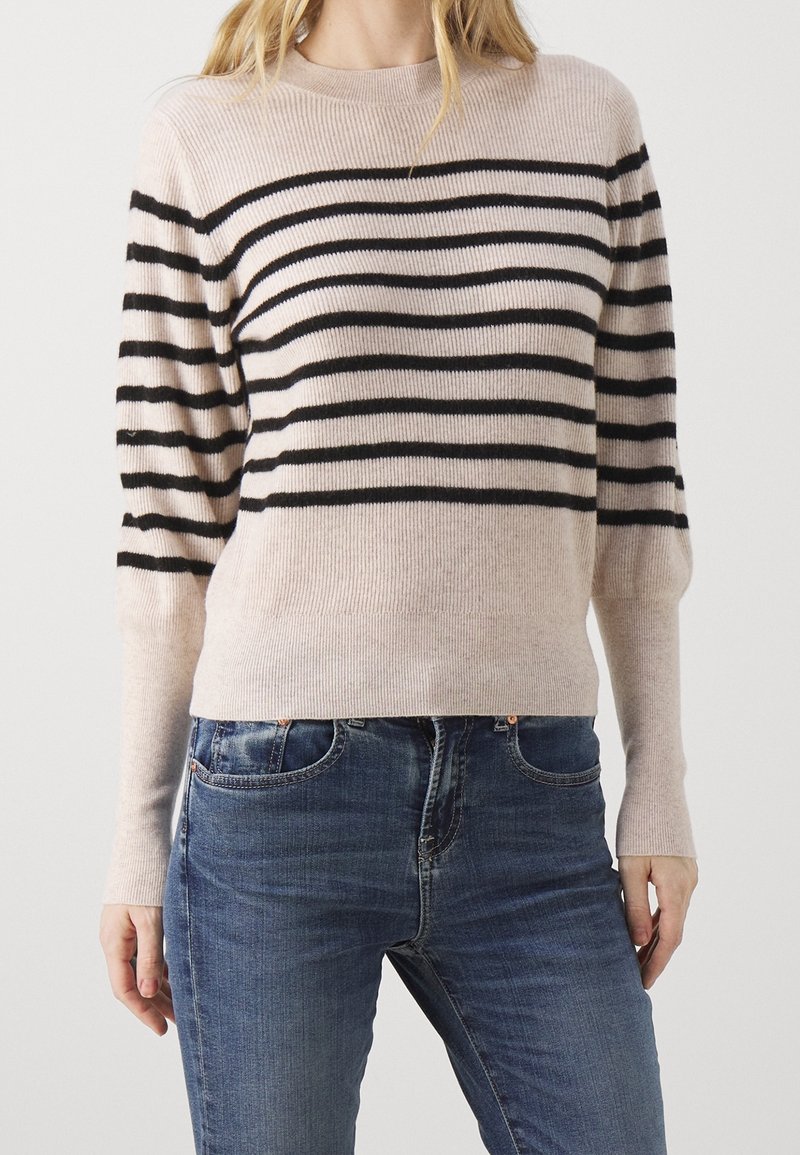Femme portant un pull côtelé beige avec des rayures horizontales noires associé à un jean en denim bleu, debout sur un fond uni.