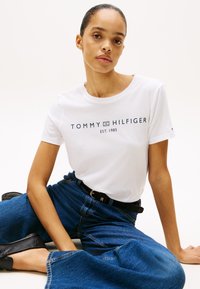 Balta medvilnės marškinėliai su tamsiai mėlynu logotipu "TOMMY HILFIGER EST. 1985." Derinami su aukštu juosmeniu mėlynais džinsiniais platėjančiais džinsais. Juodas diržas ir batai.