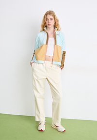 Jachetă cu fermoar de culoare bleu deschis și bej, cu accente maro, asortată cu pantaloni largi crem și adidași multicolori. Material neted cu buzunare.