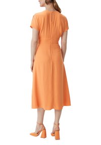Pomarańczowa sukienka midi z krótkimi rękawami, otworem w plecach i dopasowanym stanem. Połączona z pomarańczowymi sandałami na słupku. Gładka struktura materiału.