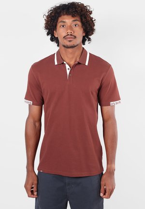Homme aux cheveux bouclés portant un polo marron à manches courtes avec des accents blancs au col et un pantalon noir, regardant devant lui avec une expression neutre.