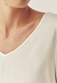 Weißes V-Ausschnitt-Oberteil aus einem glatten, leichten Stoff. Es hat einen subtilen Glanz und eine saubere Verarbeitung entlang des Hals- und Schulterbereichs.