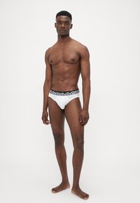 Weiße Herrenunterhosen mit schwarzem Logo-Bund, enganliegendes Design, glattes Material und minimale Abdeckung, getragen von einem stehenden Modell.