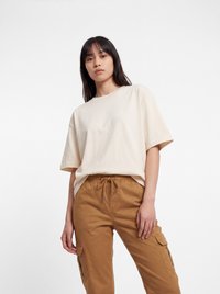 Giovane donna con lunghi capelli scuri che indossa una maglietta beige larga e pantaloni cargo marrone chiaro, in piedi davanti a uno sfondo bianco.