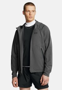 Veste zippée grise avec capuche, fabriquée en tissu lisse. Comprend deux poches latérales et un logo noir sur le côté supérieur gauche de la poitrine.
