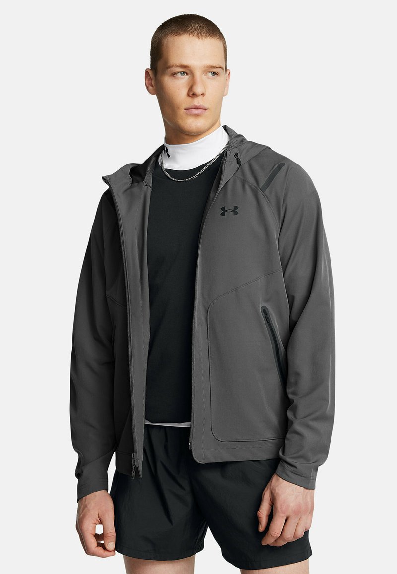 Veste zippée grise avec capuche, fabriquée en tissu lisse. Comprend deux poches latérales et un logo noir sur le côté supérieur gauche de la poitrine.