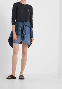 Musta pitkähihainen paita, jossa pieni logo, yhdistetty farkkuhameeseen, joka on solmittu vyötäröltä, siinä on rispaantunut helma ja kaksi etutaskua.