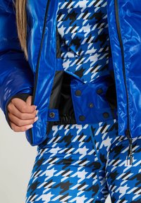 Blå glänsande puffjacka med svart foder och tryckknappar över en blå och svart houndstooth-mönstrad outfit. Närbild på tygdetaljer.
