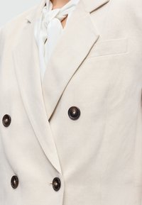 Μπεζ σακάκι με διπλό πέτο και σκουρόχρωμα κουμπιά, φορεμένο πάνω από ένα λευκό πουκάμισο δεμένο με φιόγκο στο γιακά.