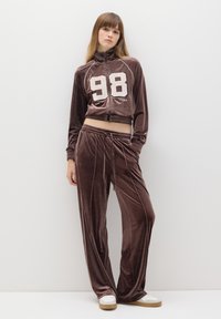 Completo in velluto marrone composto da giacca cropped con zip e il numero bianco "98", abbinato a pantaloni. I pantaloni hanno una cintura elastica e tasche laterali.