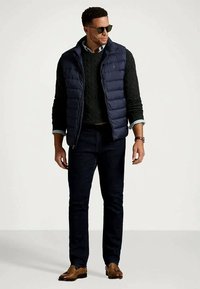 Mann mit Sonnenbrille, marineblauer Steppweste, schwarzem Zopfmusterpullover, dunklen Jeans und braunen Lederloafers, der vor einem schlichten Hintergrund steht.