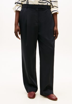 Pantaloni su misura blu navy scuro con vita alta, design a gamba larga e pieghe frontali. Il tessuto appare liscio, abbinato a scarpe slip-on color bordeaux.