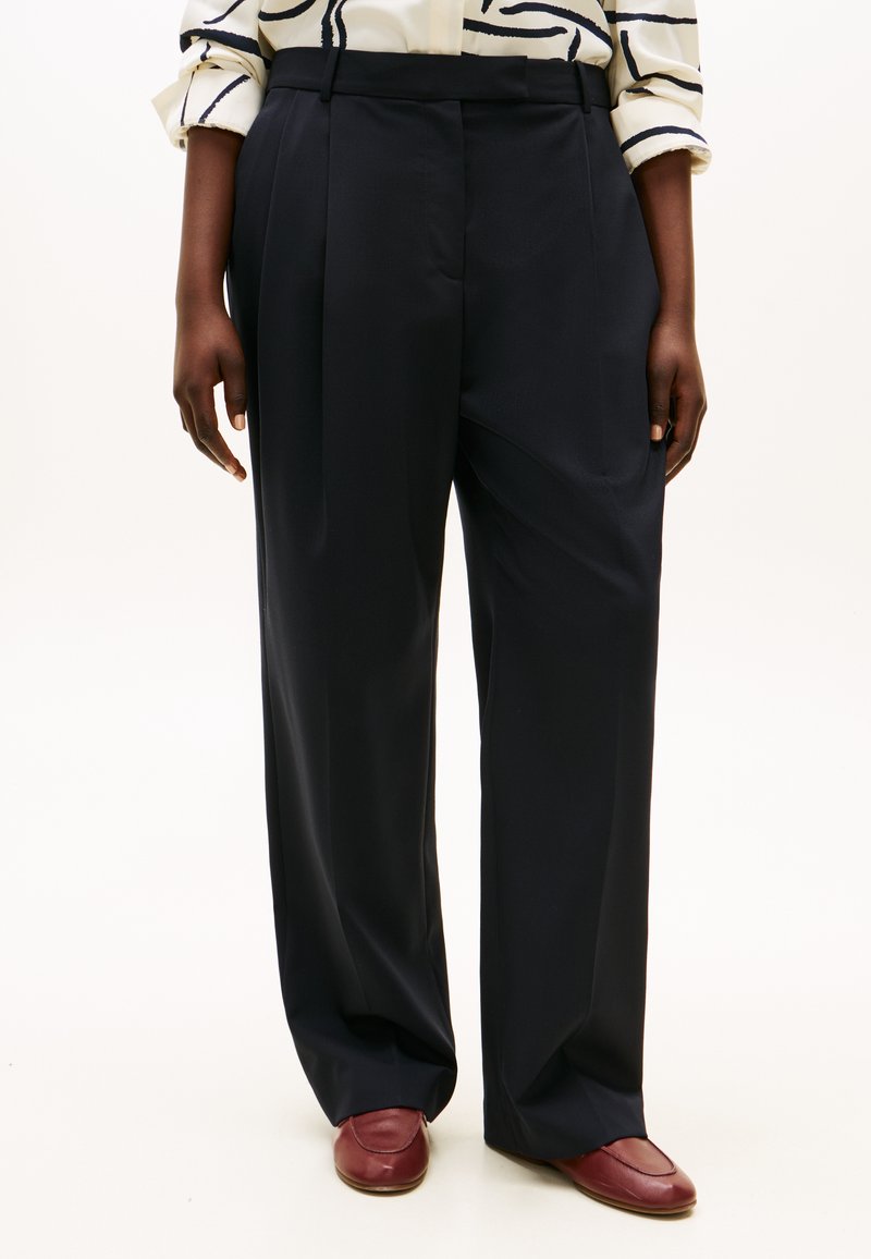 Σκούρο μπλε παντελόνι σε στενή γραμμή με ψηλή μέση, φαρδιά γραμμή και πιέτες μπροστά. Το ύφασμα φαίνεται λείο, συνδυασμένο με μπορντό παντόφλες.