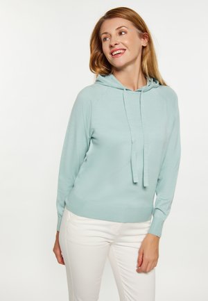 Junge Frau mit hellblauem Kapuzenpullover und weißen Hosen lächelt und schaut zur Seite vor einfachem weißem Hintergrund.