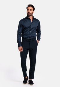 Marineblauwe shirt met lange mouwen en een glanzende afwerking, met knoopsluiting aan de voorkant, gecombineerd met donkere broek en zwarte instappers op een effen achtergrond.