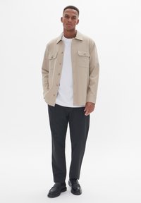 Chaqueta beige de botón con dos bolsillos en el pecho, llevada sobre una camiseta blanca y pantalones negros. La chaqueta presenta una textura suave y un brillo sutil.