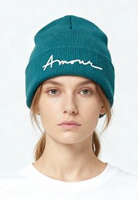 Teal gebreide muts met een afgeronde vorm en een licht opgerolde rand, met witte cursieve tekst "Amour" aan de voorkant.