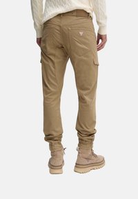 Pantalones cargo color caqui con puños elásticos, bolsillos traseros y parche con el logo. El tejido es suave con una textura ligeramente brillante. Se combinan con botas beige.