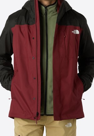 Personne portant une veste zippée bordeaux et noire sur une polaire verte avec un pantalon beige, tous arborant les logos de The North Face.