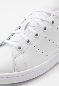 Vit lädersneaker med rund tå, perforerade detaljer, platta vita snörningar och en texturerad gummisula. Minimalistisk design.