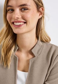 Giacca di un beige chiaro, con texture e colletto arrotondato indossata sopra una maglietta bianca. Vista ravvicinata di una persona sorridente. I capelli naturali ricadono da un lato.