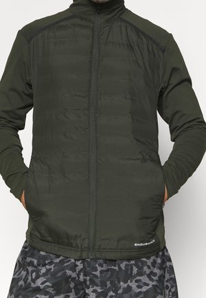 Mand iført en mørkegrøn lynlåstrøje med hænderne i lommerne og sorte-grå camouflage shorts, stående foran en neutral baggrund.