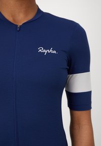 Marineblå sykkeltrøye med glidelås foran, strukturert stoff, korte ermer og en hvit horisontal stripe på overarmen med "Rapha"-logo.