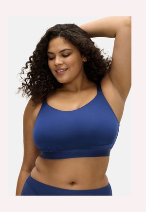 Femme aux cheveux bouclés souriante, un bras levé, portant un soutien-gorge de sport bleu et une culotte assortie sur fond blanc.