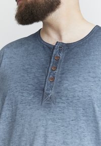 Blå, tekstureret kortærmet skjorte med rund halsudskæring, der har en knapstolpe med fire brune knapper og kontrasterende syninger.