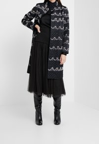 Abrigo negro acolchado con cuentas decorativas y bordado, con mangas largas y cuello alto, combinado con una falda de tul negra y botas hasta la rodilla.