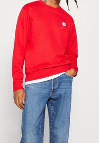 Sudadera roja con cuello redondo y puños acanalados, con un pequeño parche de logo azul. Se lleva con vaqueros azules de mezclilla.