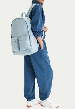 Persona con chándal azul y zapatillas blancas lleva una mochila azul claro con bolsillos delanteros y logo de Reebok.