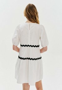 Biele šaty s nafúknutými rukávmi, so čiernym rickrack lemovaním v pase. Na chrbte je malý otvor v tvare kľúča a čistý šev.