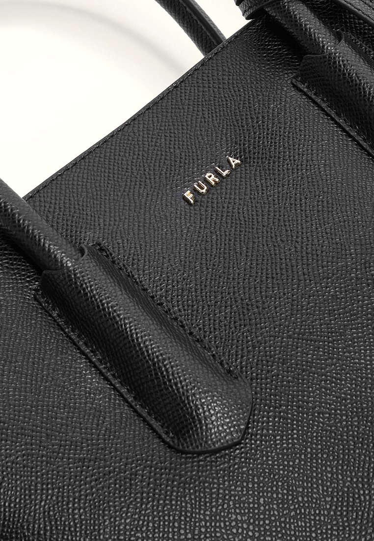 Bolso de mano de cuero negro texturizado con dos asas resistentes, que presenta un logotipo "FURLA" en tono dorado en la parte frontal. Patrón de grano sutil en toda la superficie.