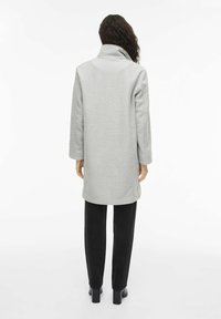 Gaiši pelēks mētelis ar augstu apkakli, garām piedurknēm un brīvu piegriezumu, kopā ar melnām biksēm un zābakiem ar papēdi. Teksturēts auduma apdare.